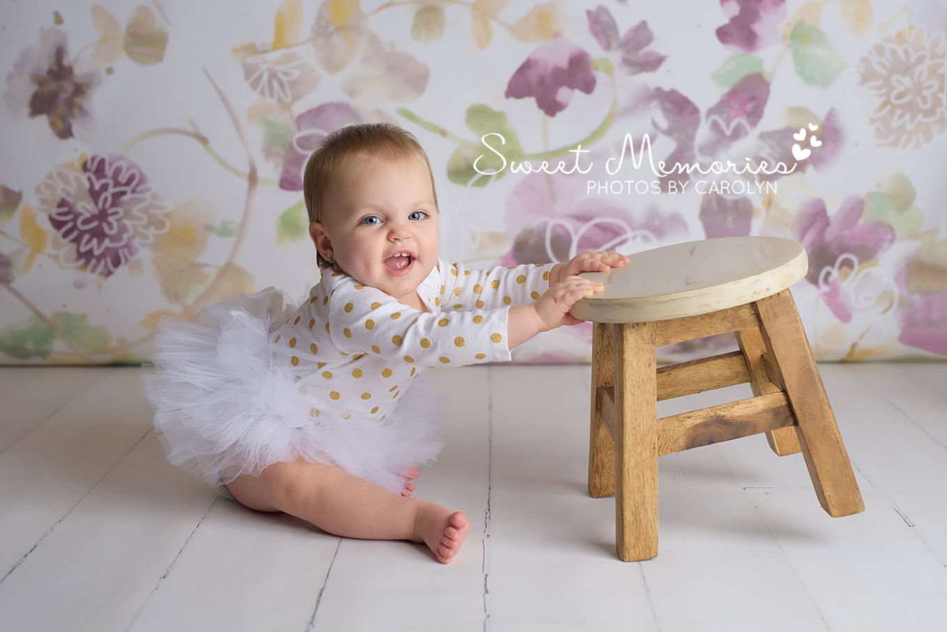 Purple and gold floral backdrop with smiling one year old girl in a tutu with stool in Quakertown, PA | Sweet Memories Photos by Carolyn