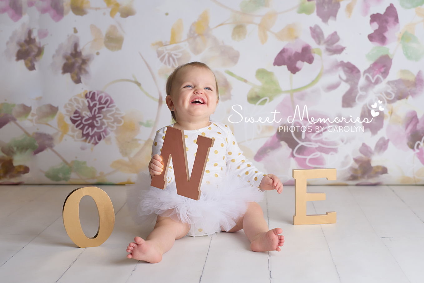 Purple and gold floral backdrop with smiling one year old girl in a tutu with ONE letters in Quakertown, PA | Sweet Memories Photos by Carolyn | First Birthday Baby Photographer