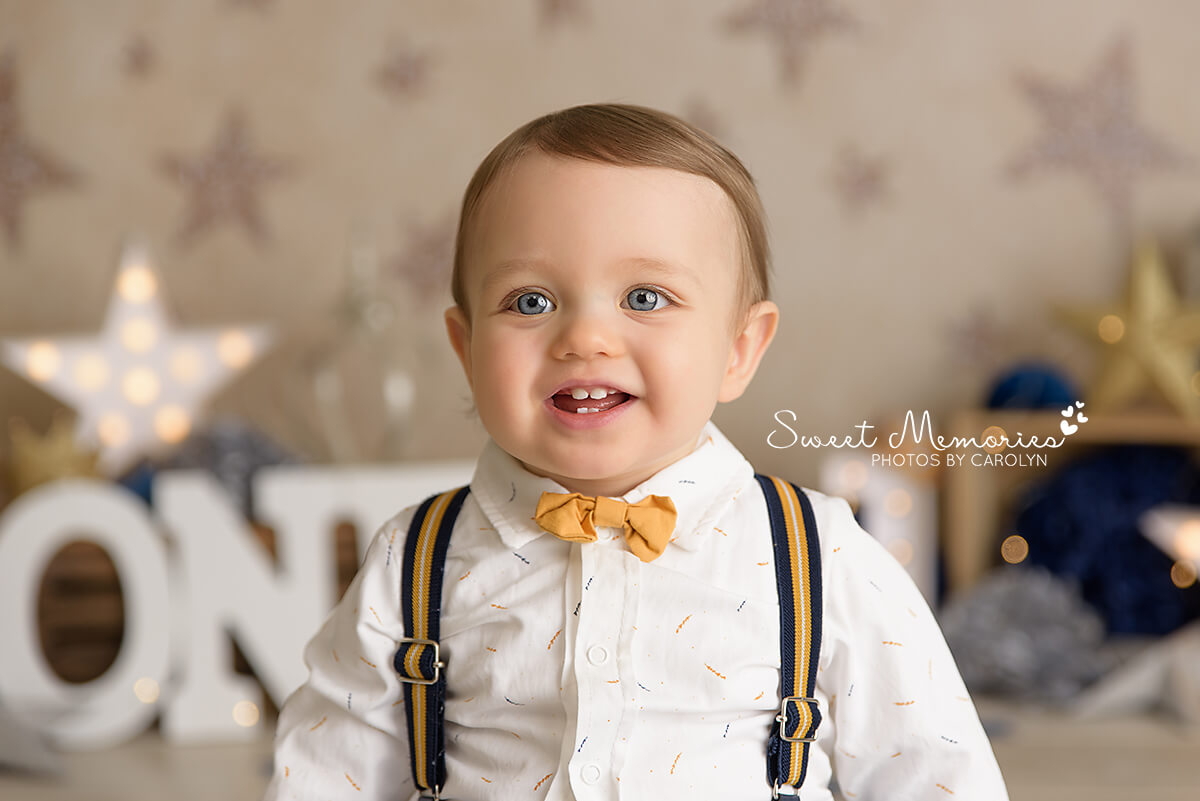 close up of one year old baby boy in yellow suspenders and bow tie smiling with star backdrop