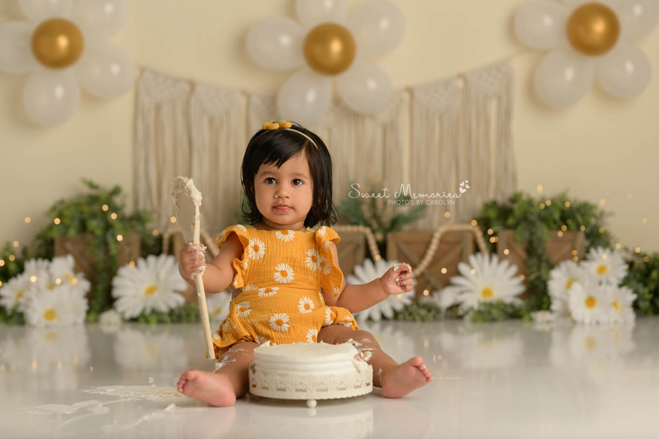 one year old baby girl holding spoon with cake with boho daisy backdrop | First birthday cake smash photography Round Rock TX