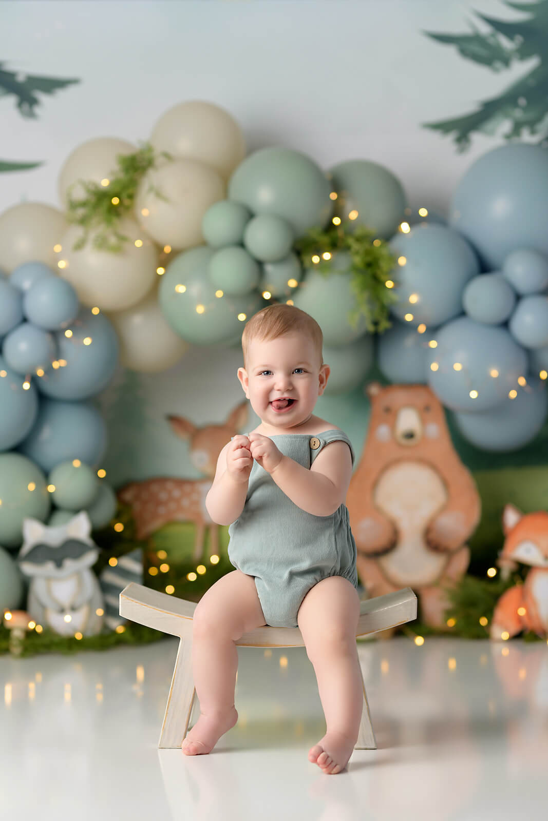 Baby boy in blue romper smiling and sitting on a curved bench. Background is blue and green balloons on with forest animals